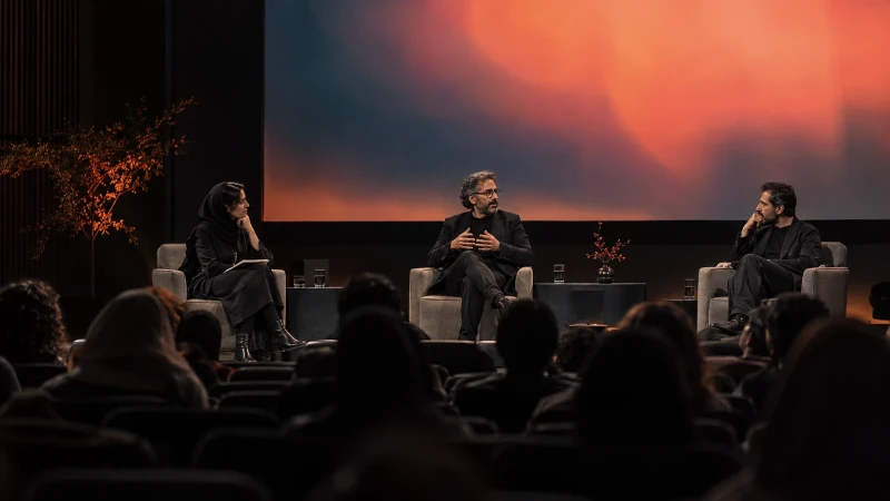 Three speakers on a panel stage with a lit projection screen behind them
